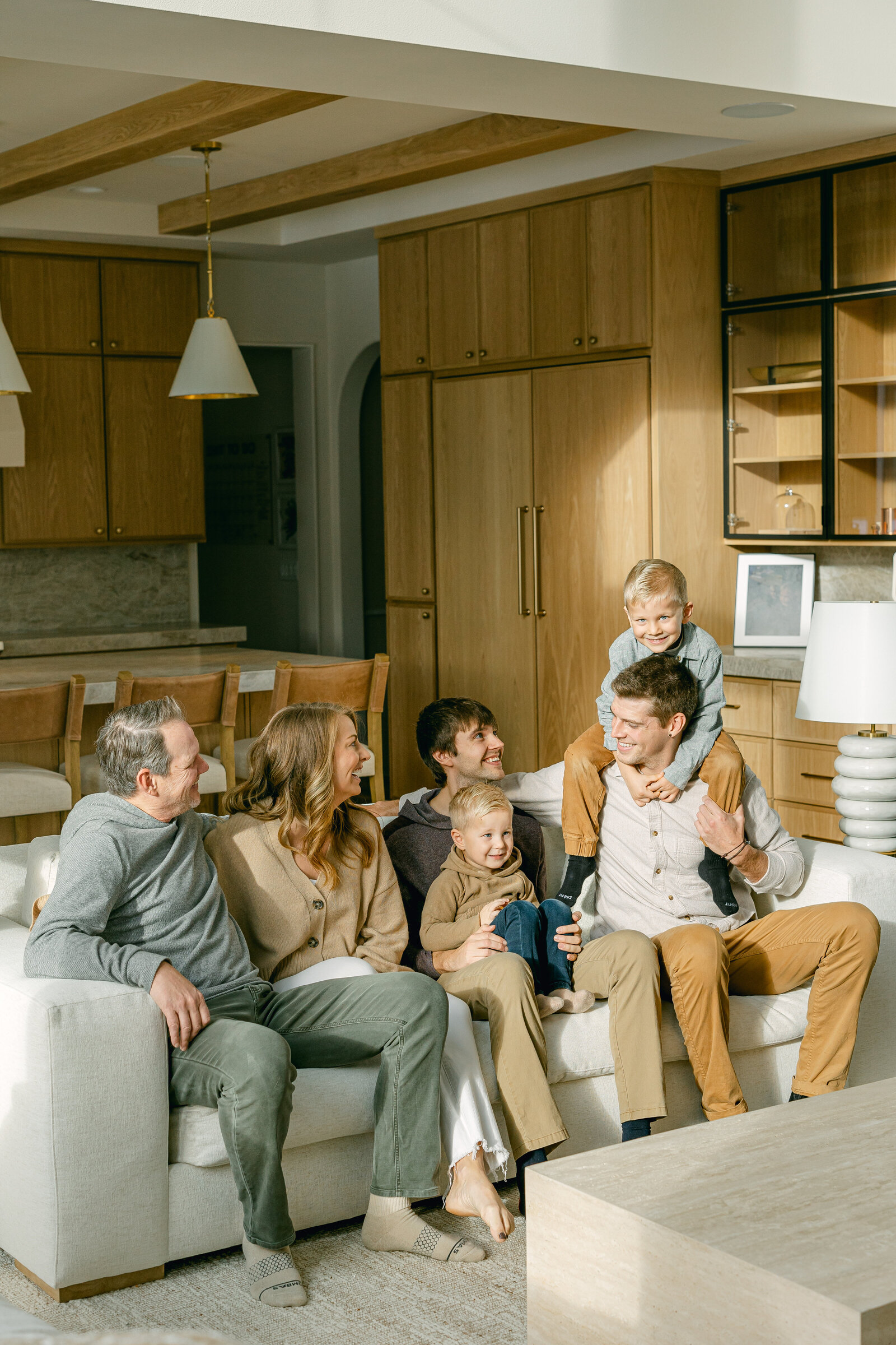 Six family members including two young boys sit laughing together on a white sofa in a living room.