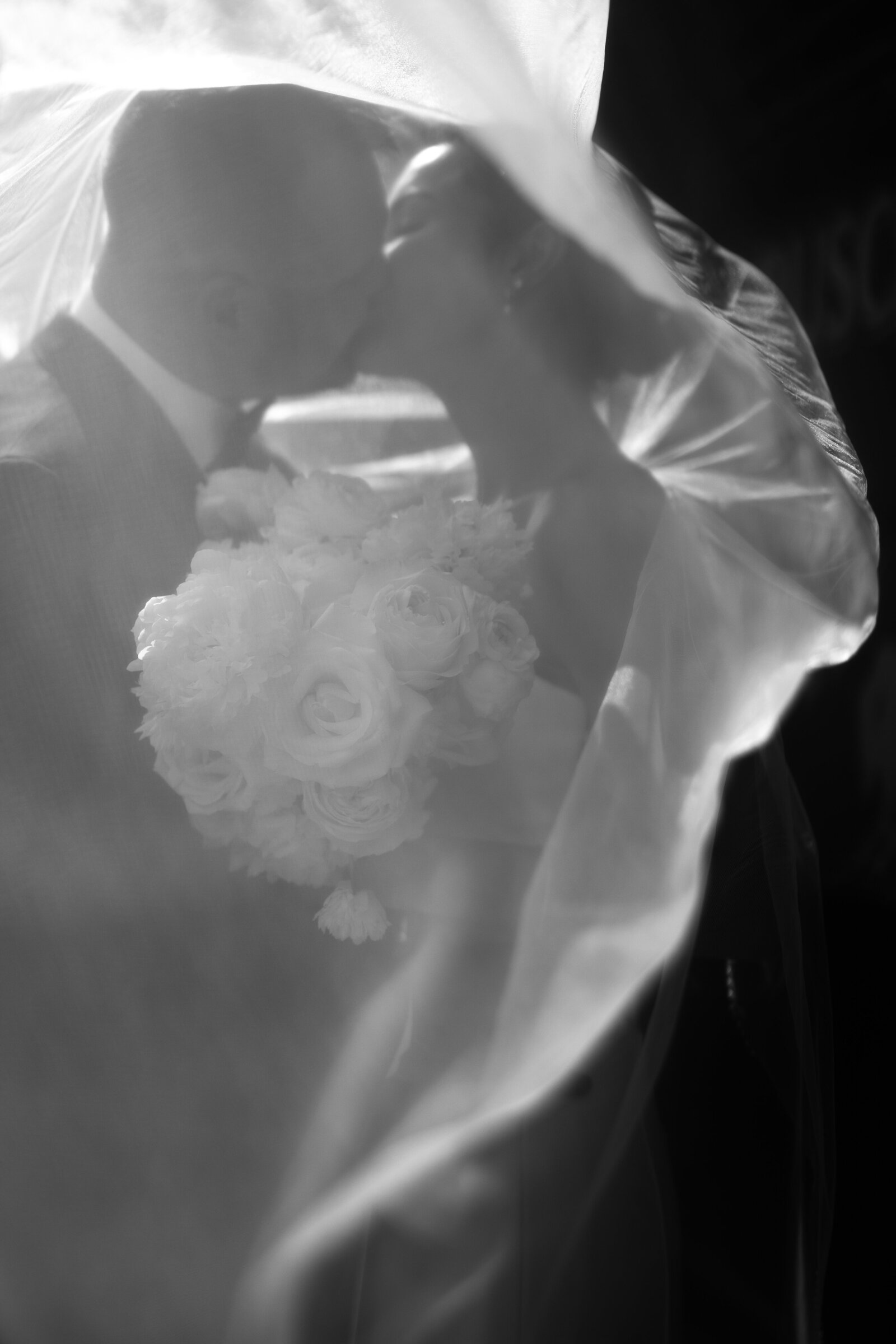Black and white photo of a couple kissing beneath a veil above a white bouquet.